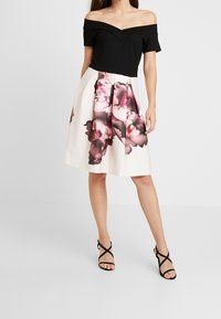 Svart off-shoulder topp i kombination med en vit kjol som har ett stort rosa blommönster. Modell bär svarta sandaler med remmar.