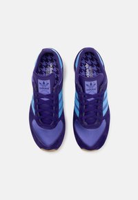 Paire de baskets Adidas violettes avec des rayures bleu clair et des semelles intérieures à motifs, vues de dessus sur fond blanc.
