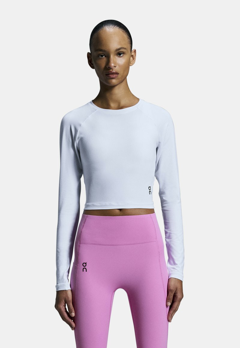 Weißes Langarm-Crop-Top mit Raglanärmeln, kombiniert mit hochtaillierten rosa Leggings, die ein Logo-Detail am linken Oberschenkel aufweisen.