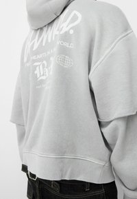 Felpa grigia oversize con cappuccio regolabile, caratterizzata da grafiche bianche stampate sul retro e bordi grezzi sulle maniche.