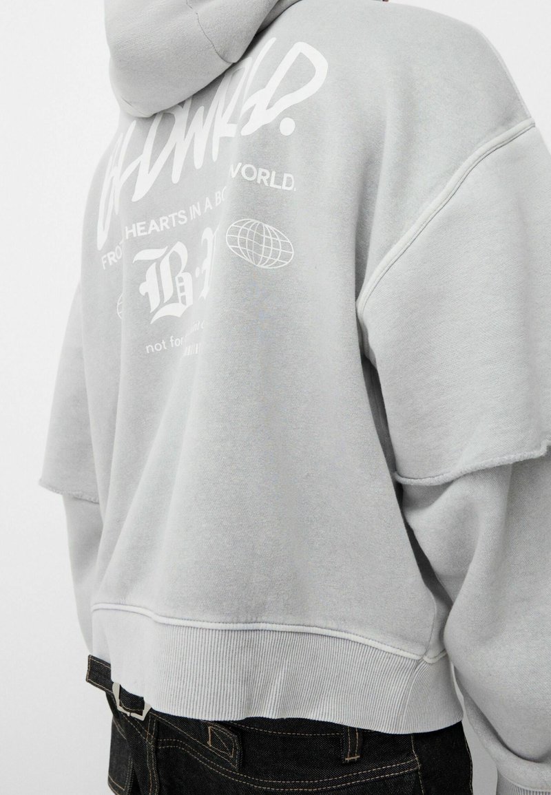 Felpa grigia oversize con cappuccio regolabile, caratterizzata da grafiche bianche stampate sul retro e bordi grezzi sulle maniche.