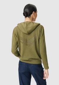 Un sweat à capuche vert olive avec un motif chevron texturé au dos, doté d'une capuche avec cordon de serrage et de poignets côtelés, associé à un jean bleu foncé.
