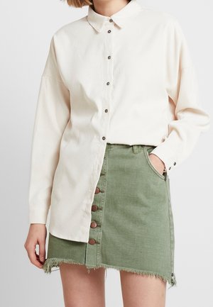 Kobieta ubrana w kremową koszulę z długimi rękawami zapinaną na guziki oraz zieloną spódnicę dżinsową na guziki z postrzępionym dołem.