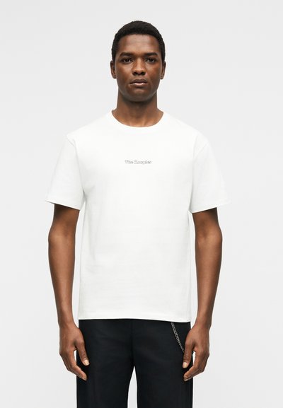 Άνδρας που φοράει απλό λευκό κοντομάνικο μπλουζάκι με μικρό κεντρικό κείμενο "The Kooples", σε συνδυασμό με μαύρο παντελόνι και ασημένιο αξεσουάρ αλυσίδας.