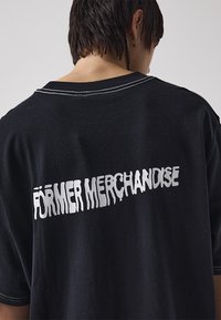 Black oversized t-shirt featuring white faded "FÖRMER MERCHANDISE" text on the back, with contrasting stitching details along the neckline.