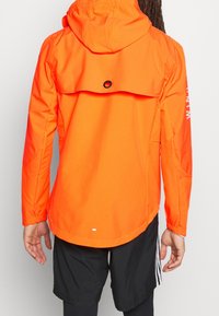 Giacca leggera arancione con cappuccio, presenta polsini elasticizzati, una cornetta di ventilazione e una varietà di texture nel tessuto.