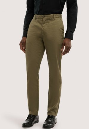 Mężczyzna noszący oliwkowozielone, dopasowane chinosy z przednimi kieszeniami, w połączeniu z czarnymi eleganckimi butami i czarną koszulą z długim rękawem.