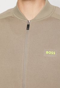 Beige bunda na zip s ribovanou texturou, s vysokým límcem a výrazným zeleným logem "BOSS" na levé přední straně.