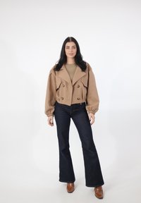 Gekürztes, zweireihiges Jackett aus hellbraunem Wollmischgewebe mit breiten Revers und überdimensionalen Ärmeln, kombiniert mit dunklen, ausgestellten Jeans und braunen Stiefeletten.