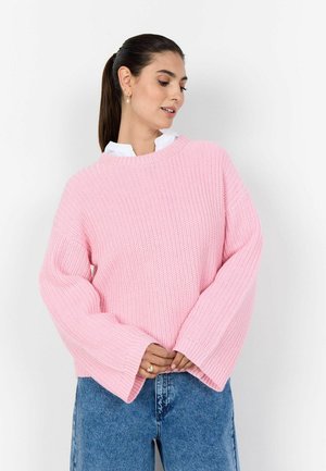 Vrouw met een donkere paardenstaart, goudkleurige oorbellen, een roze oversized gebreide trui over een wit shirt en een blauwe spijkerbroek, kijkt omlaag en houdt haar trui vast.