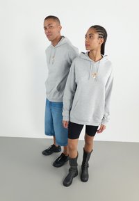 Hanorace gri cu glugă și buzunare frontale, având un mic design pe piept. Modelul este îmbrăcat cu pantaloni scurți din denim și cizme negre.