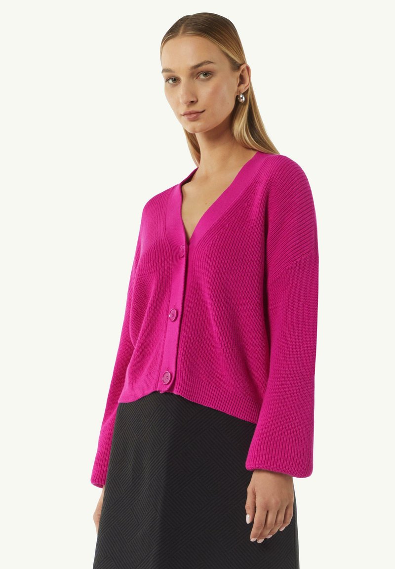Fuchsia Strickjacke mit V-Ausschnitt, Knopfverschluss, gerippter Textur und breiten Ärmeln. Kombiniert mit einem strukturierten schwarzen Rock.