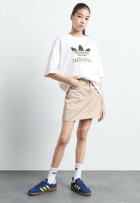 T-shirt oversized branca com o logo verde da Adidas, saia bege, sapatilhas azuis com detalhes em neon e meias brancas com o logo da Adidas.