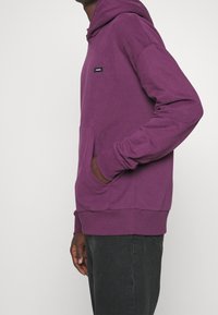 Paarse hoodie met een voorzak, ribbelmanchetten en onderkant. Heeft een klein zwart logopatch op de borst. Zacht, gestructureerde stof.