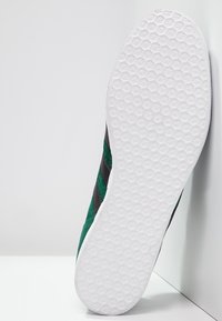 Gröna mockaskor med svarta diagonala ränder, vit strukturerad gummisula och sexkantigt mönster på yttersulan.