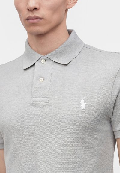 Gros plan d'une personne portant un polo gris clair avec deux boutons et un petit logo blanc brodé sur la poitrine.