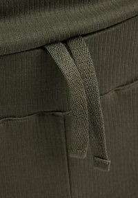 Olivgrüner, gerippter Stoff mit weicher Textur, der am Taillenbund Kordeln als Verschluss aufweist. Das Material wirkt dehnbar und langlebig.