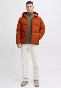 Padded orange jacket with a hood, zip front, and elastic cuffs paired with a green sweater, white pants, and beige shoes on a neutral background.