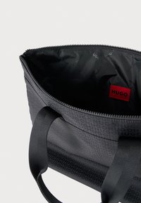 Čierna taška tote s textúrovaným vonkajškom, na ktorom sa opakuje logo 'HUGO', so zipsovým uzáverom a pevnými textilnými rúčkami. Vnútro je hladké čierne.