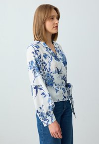 Weiße Bluse mit floralen Mustern in Blau von Blumen und Vögeln, langärmelig und mit einem Wickeldesign, das mit einem Band in der Taille fixiert ist.