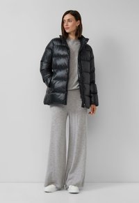 Melna pūkaina jaka ar izšūtu dizainu, augstu apkakli un ar rāvējslēdzēju valkāta priekša, kombinēta ar gaiši pelēku džemperi un platām biksēm.