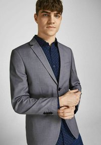 Grå skræddersyet blazer med en enkelt sort knap, båret over en mørkeblå skjorte med hvide prikker. Glat tekstur, struktureret pasform.
