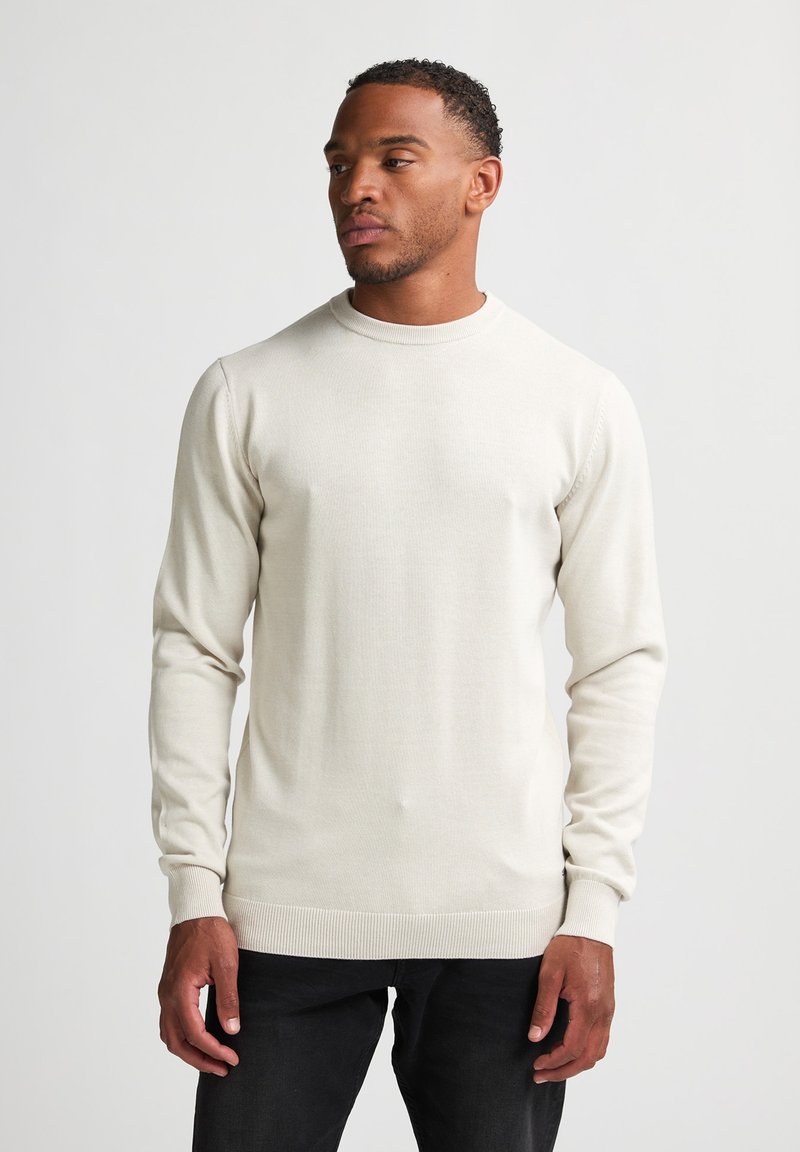 Pulover bej deschis cu guler rotund, fabricat dintr-un material tricotat texturat, având mâneci lungi și manșete și tiv din jerseu. Purtați cu blugi închis la culoare.