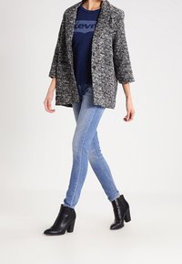 Blazer gris texturizado sobre una camiseta azul marino con un logo, combinado con jeans ajustados azul claro y botas de tobillo negras con un tacón grueso.