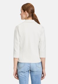Biała marynarka o dopasowanym kroju z rękawami do łokcia, z teksturowanej tkaniny, z wyraźnie zarysowanym fasonem, w połączeniu z jasnoniebieskimi dżinsami.