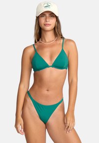 Set bikini in turchese con spalline sottili e coppe triangolari. Il pantalone presenta uno stile a copertura minima. Indossato con un cappello beige e una collana a strati.
