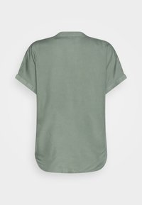 Bakvy av en enkel kortärmad skjorta i salviagrön med uppvikta ärmar och rundad nederkant på en vit bakgrund.