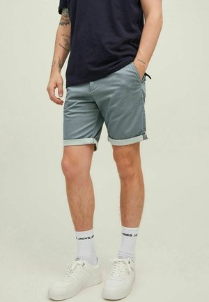 Mężczyzna ubrany w podwinięte szare spodenki, czarną koszulkę, białe skarpetki z napisem "JACK & JONES" oraz białe trampki, stojący z jedną ręką w kieszeni.