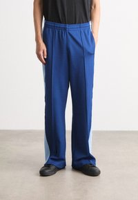 Pantalones deportivos azules con una textura suave, que presentan paneles laterales en azul claro. Cintura elástica con bolsillos, combinados con zapatos negros.
