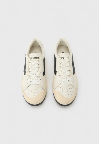 Paire de baskets en cuir blanc avec des bandes latérales noires, des bouts de chaussures crème, des détails perforés et des lacets blancs sur un fond gris clair.