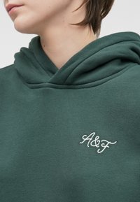Green hooded sweatshirt made of soft fabric. Features an embroidered white "A&F" logo on the front. Hood has a relaxed, draped design.