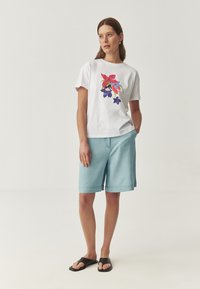 Hvid bomulds T-shirt med farverigt blomsterprint, parret med lyseblå shorts og sorte slidesandaler. Enkel, afslappet pasform.
