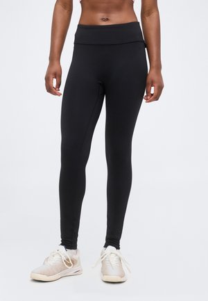 Personne portant des leggings noirs ajustés et des chaussures de sport blanches, debout devant un fond clair uni.