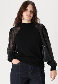 Μαύρος πουλόβερ με ψηλό λαιμό και διαφάνεις παγιέτες στα μανίκια, συνδυασμένο με σκούρα μπλε τζιν και μία μικρή μαύρη τσάντα.