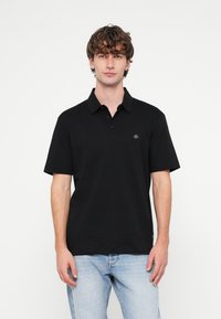 Melnā polo krekls ar klasisku apkakli, īsām piedurknēm un nelielu logotipu uz krūtīm. Izgatavots no mīksta auduma, ar brīvu piegriezumu.