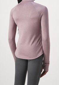 Kvinde set fra ryggen iført en mauve langærmet tætsiddende træningstrøje med tommelfingerhul og mørkegrå leggings.