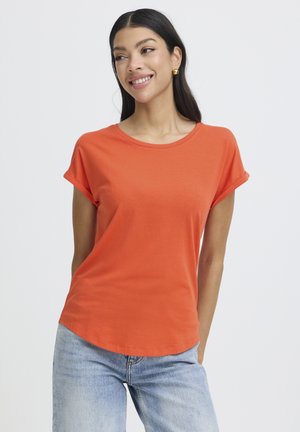 Kobieta o długich ciemnych włosach uśmiecha się, ubrana w pomarańczowy T-shirt z krótkim rękawem i jasnoniebieskie dżinsy, na tle jednolitego jasnego tła.