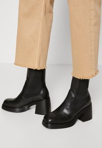 Botas negras hasta el tobillo con un tacón grueso, hechas de cuero brillante. Presentan paneles elásticos en los lados y punteras redondeadas. Combinadas con pantalones beige recortados y deshilachados.