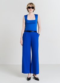 Top bleu sans manches avec un décolleté carré, associé à un pantalon bleu à jambes larges et des mules noires à talons ornées de clous décoratifs.
