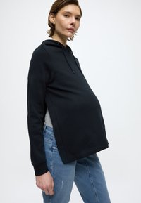 Μαύρο φούτερ με κουκούλα και άνετη εφαρμογή, κατασκευασμένο από μαλακό υλικό, με πλευρικές σχισμές, κουκούλα με κορδόνι, συνδυασμένο με ανοιχτόχρωμο μπλε τζιν.