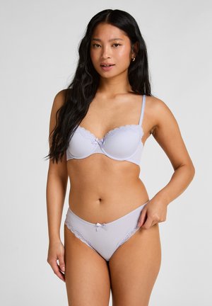 Vrouw met lang donker haar, gekleed in een lila geribbelde beha en bijpassende slip met kantafwerking, staand tegen een effen lichte achtergrond.