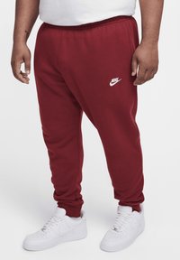 Red sweatpants with a drawstring waist, featuring a white Nike logo on the left thigh and cuffs at the ankles, paired with white sneakers.
