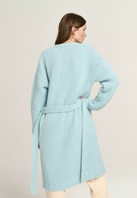 Long, light blue knitted cardigan with a wrap tie at the waist, featuring a relaxed fit and ribbed cuffs. Soft, textured fabric.