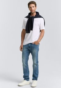 Vit T-shirt med blå logotyp, blå denimjeans, marinblå tröja knuten runt nacken och vita sneakers. Avslappnad outfit med enkel design.