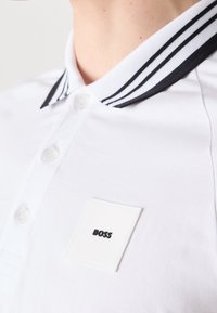 Polo blanc avec col rayé noir et petit écusson carré portant l'inscription "BOSS" sur la poitrine, porté par une personne.