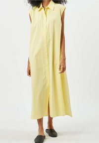 Abito midi giallo senza maniche, con bottoni e colletto, indossato con scarpe slip-on nere, modello in piedi su uno sfondo bianco semplice.